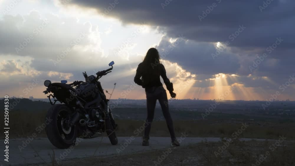 Young biker woman silhouette  dancing on cloudy sunset near her Motorcycle on the desert road when Airplane flying in sky
