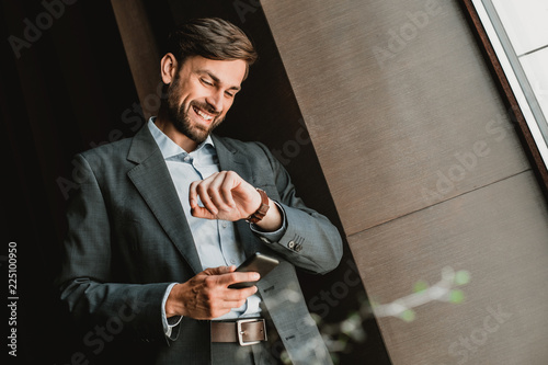 Low angle of smiling businessman wearing suit and standing indoor. He is holding mobile phone and checking time on wrist