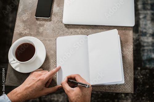 Top view of coffee table with business appliances and mug with hot tea. Man is writing information with pen in diary while using laptop and smartphone