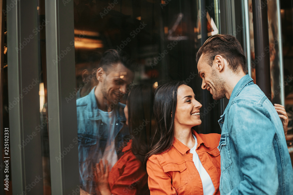 Side view smiling female hugging beaming male while leaning against window with their reflection outside. Glad couple communicating during rest concept