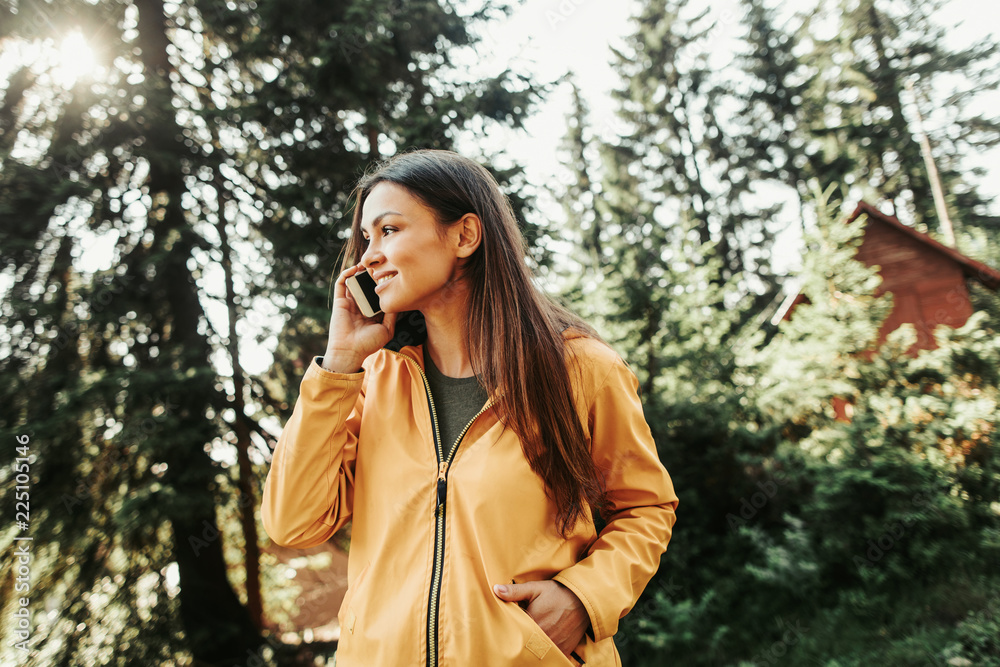 Finally i am chatting with you. Waist up portrait of beautiful young lady enjoying phone conversation while standing in sunny wood. She is looking away with nice smile and keeping hand in pocket Finally i am chatting with you. Waist up portrait of beautiful young lady enjoying phone conversation while standing in sunny wood. She is looking away with nice smile and keeping hand in pocket