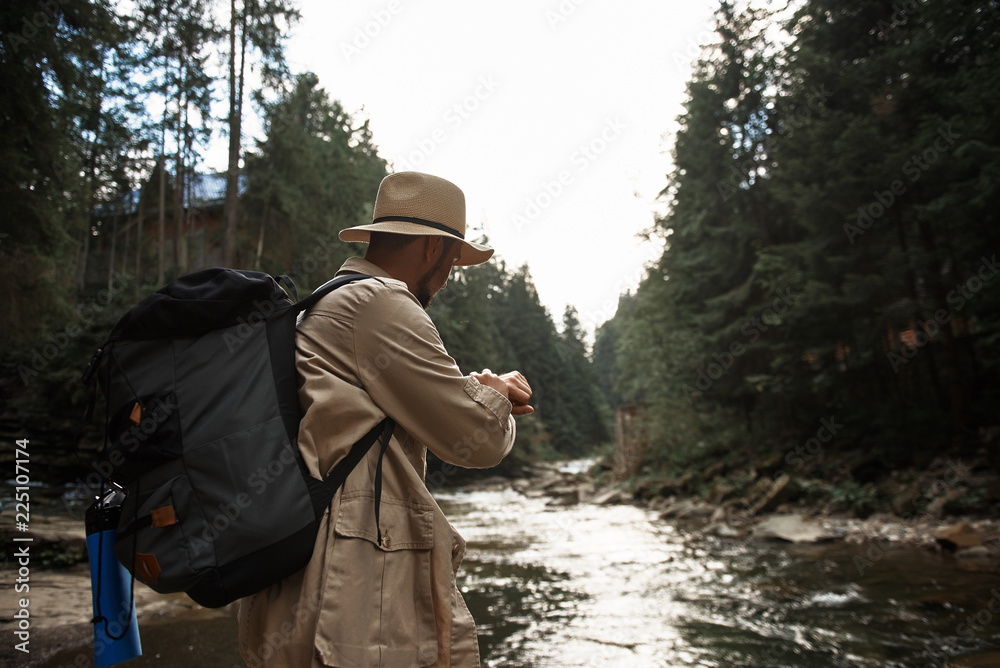 Checking time. Confident experienced traveler carrying his backpack and looking at the watch while standing next to the mountain river Checking time. Confident experienced traveler carrying his backpack and looking at the watch while standing next to the mountain river