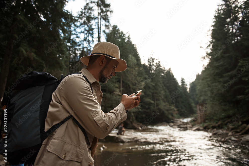 Worried traveler. Calm concentrated young man standing next to the river with big backpack and thoughtfully looking at the screen of his modern device