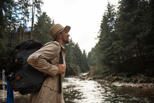Inspired by journey. Optimistic bearded young man carrying big backpack and looking confident while standing next to the mountain stream and smiling © Yakobchuk Olena