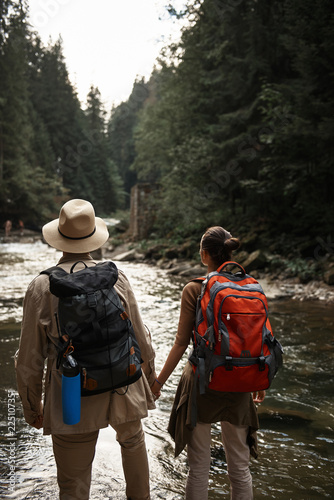 Looking at water. Calm active couple holding hands and looking into the distance while standing with big backpacks and looking at the beautiful river