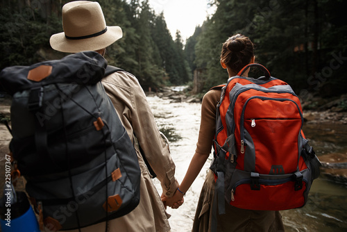 Family journey. Enthusiastic young couple of hikers carrying heavy backpacks and holding hands while looking at the mountain river © Yakobchuk Olena