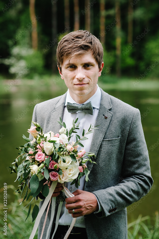 The attractive groom in a suit and bow tie, jacket, is standing and holding in hand bouquet of pink flowers and greens with ribbon at nature on the in the garden park. The attractive groom in a suit and bow tie, jacket, is standing and holding in hand bouquet of pink flowers and greens with ribbon at nature on the in the garden park.