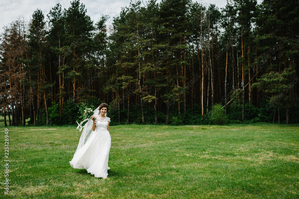 Young beautiful bride in an elegant dress is walking on the field near the forest and holding bouquet of pink flowers and greens with ribbon at nature. Outdoors. After the wedding ceremony. Young beautiful bride in an elegant dress is walking on the field near the forest and holding bouquet of pink flowers and greens with ribbon at nature. Outdoors. After the wedding ceremony.