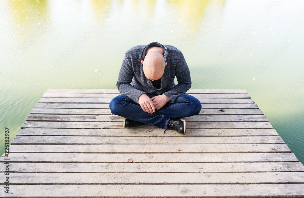 Bald young man sitting on the pier in the park and meditating Bald young man sitting on the pier in the park and meditating