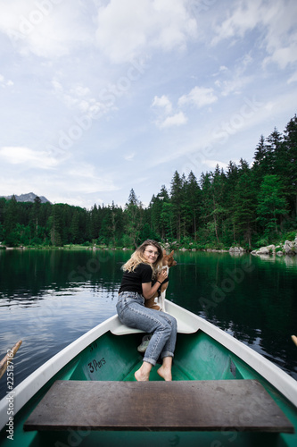 Cute, young woman or girl in denim jeans with blond hair sits in the front of vintage rowing boat floating in clean and clear waters on alpine mountain lake with her best friend puppy dog on lap © BublikHaus