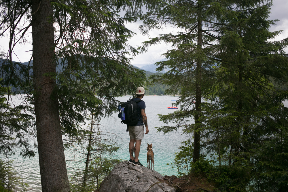 Fototapeta premium Young adventurous man with backpack and travel companion little puppy dog, best friend explore forest nature in mountains together.Stand in trees next to lake, tourism in rural aread,camping lifestyle