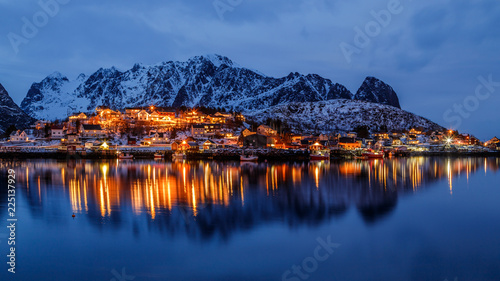 Lofoten islands, Norway. Colorful winter landscape in blue hours. Illuminated fishing village reflected in water. Snowy mountains in background.  © Telly
