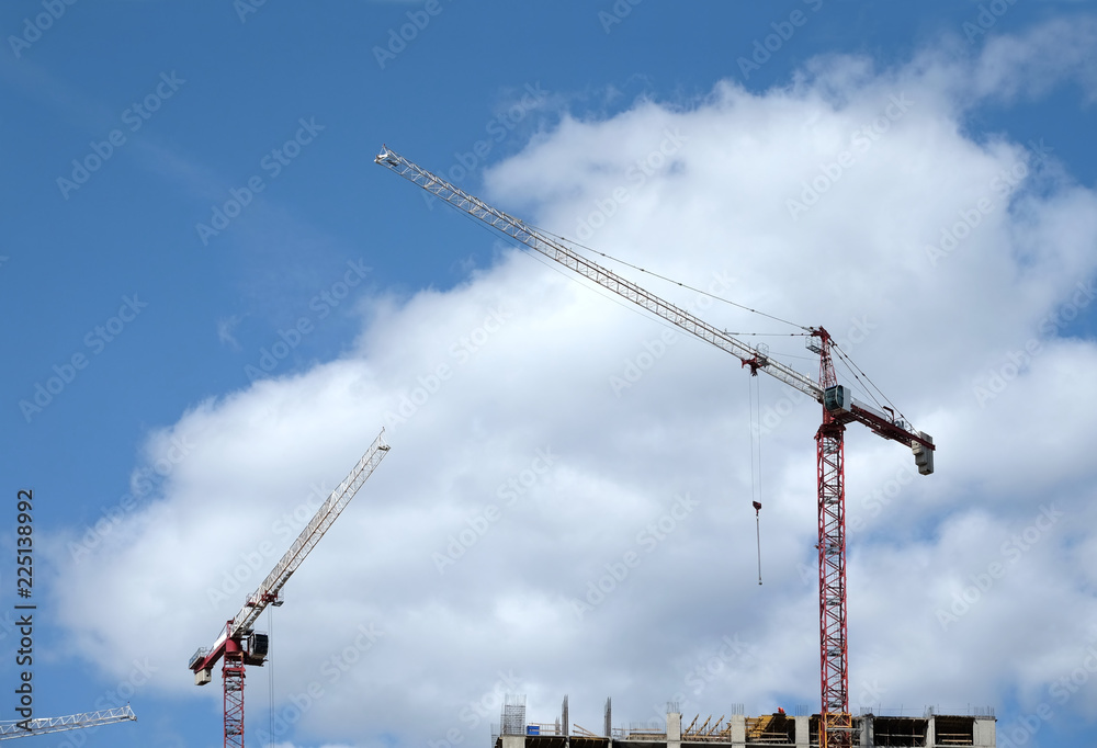 Big hoisting tower cranes and top section of modern construction building in a city over blue sky with white clouds