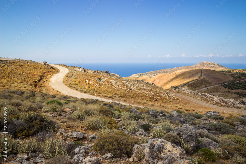 Empty road cutting through yellow hill with a sea in the distance, Crete, Greece