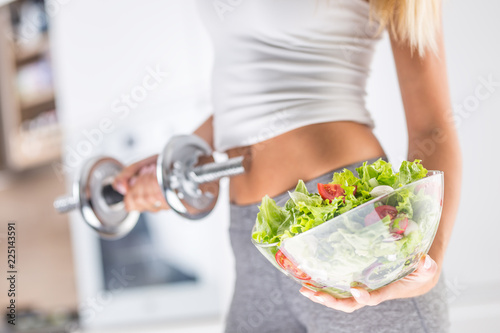 Woman holding bowl from fresh vegetable salad and dumbbell.