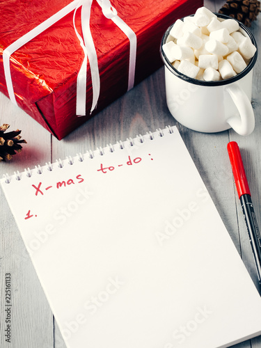 Christmas to-do list. Xmas gifts shopping planning. Make shopping or to-do list for Christmas. Notebook, mug hot chocolate with marshmallows and New Year's gift on gray wooden background.