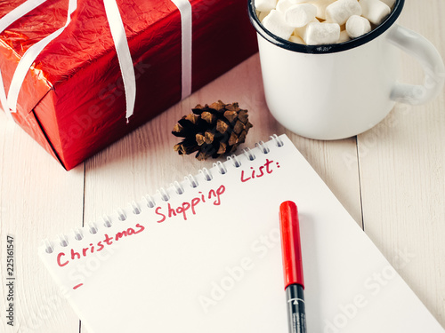 Christmas gifts shopping planning. Make shopping or to-do list for Christmas. Notebook, mug hot chocolate with marshmallows, New Year's gift and pine cone on white wooden background.