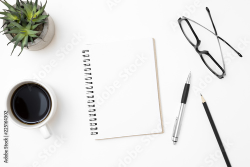 Blank notebook is on top of white office desk table with coffee cup and office supplies. Top view with copy space, flat lay.