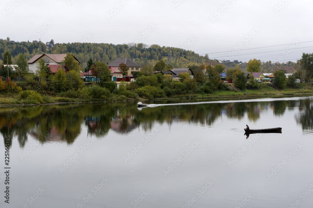 village on the bank of the autumn river in cloudy weather, in the middle of the river you can see a boat with a fisherman