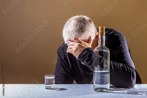 an elderly man sits at a table with a bottle of alcohol

