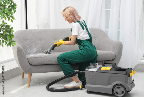 Female janitor removing dirt from sofa with upholstery cleaner in room