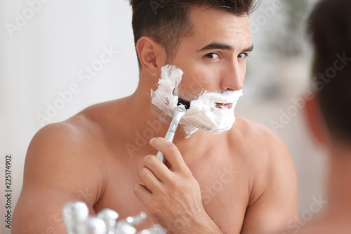 Young man shaving near mirror in bathroom