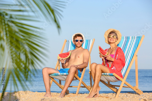 Young couple with watermelon slices in beach chairs at seacoast