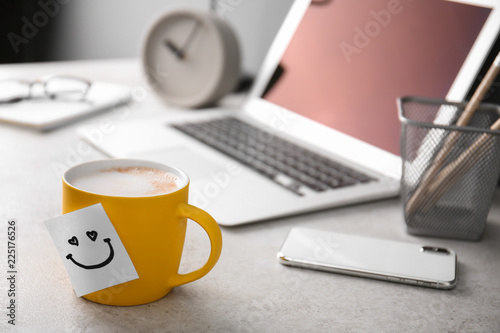 Sticky note with funny face attached to cup of coffee on office table. Space for text