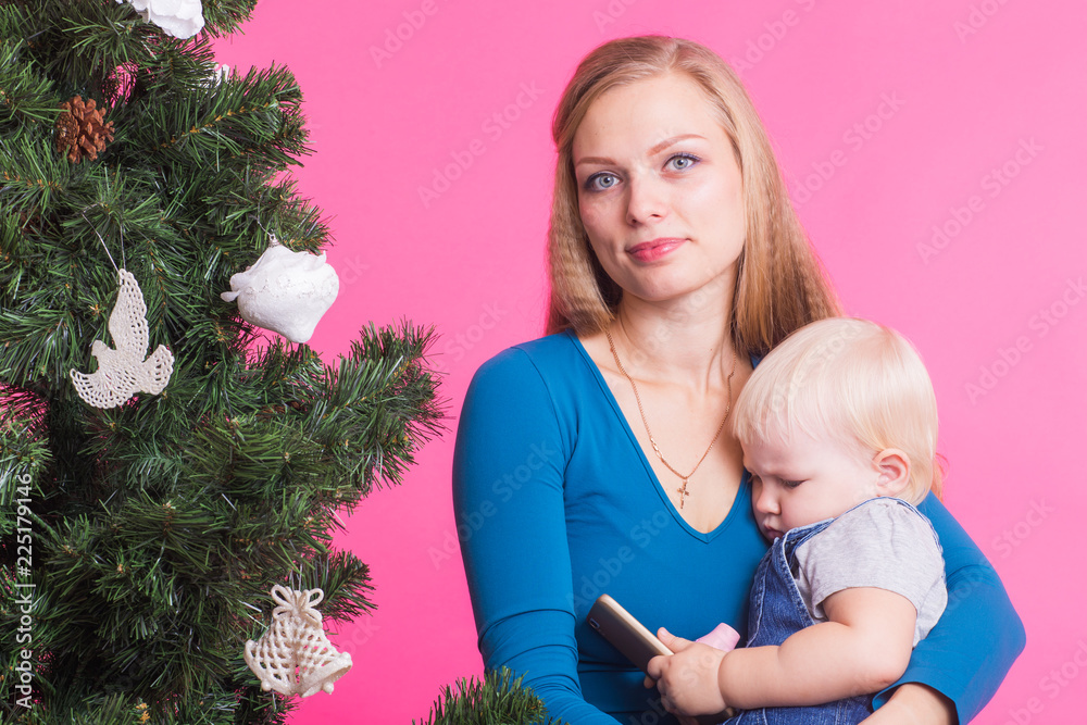 Holidays, family and christmas concept - young woman with her baby near christmas tree on pink background Holidays, family and christmas concept - young woman with her baby near christmas tree on pink background