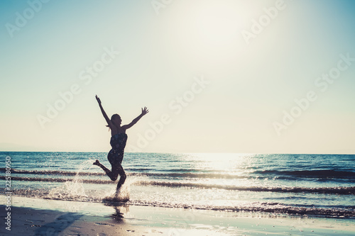 young happy girl or woman on sunset or sunrise beach running, summer vacation, sunny, positive mood, splashing water, silhouette