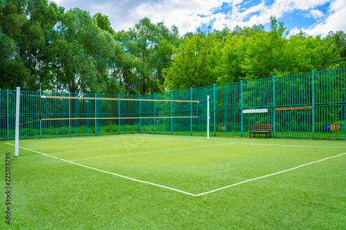 View of the sports playground in the park with artificial grass and a stretched net on a background of green trees and cloudy sky