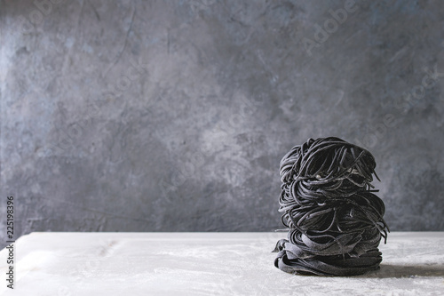 Variety of italian homemade raw uncooked cuttlefish ink black pasta spaghetti and tagliatelle in stack with semolina flour on white marble table. Copy space.