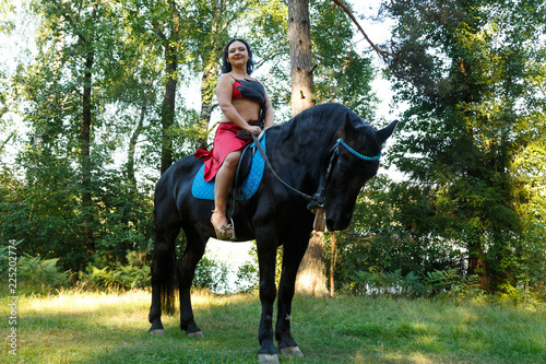 Beautiful brunette in a red suit in a saddle on a horse in the forest. Overall plan.