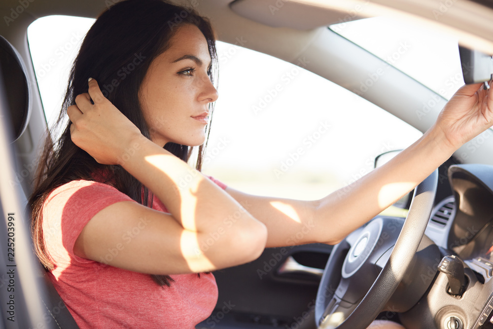 Shot of beautiful brunette woman dressed in casual t shirt, looks seriously at mirror of car, has thoughtful expression, concenrated into distance. Horizontal view of gorgeous lady in automobile Shot of beautiful brunette woman dressed in casual t shirt, looks seriously at mirror of car, has thoughtful expression, concenrated into distance. Horizontal view of gorgeous lady in automobile