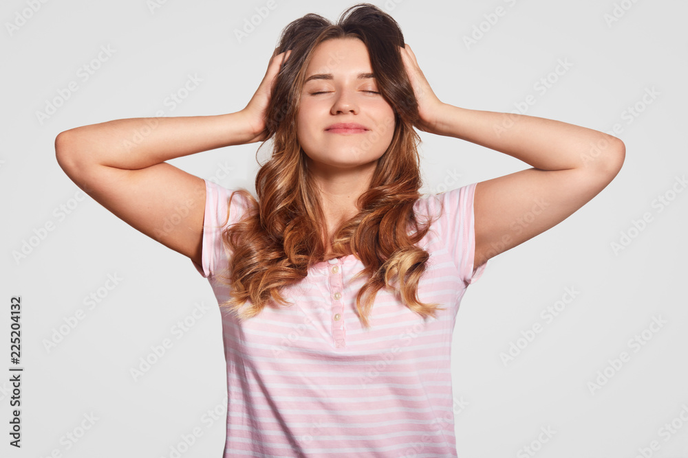 Photo of pleased realxed young woman keeps both hands on head, eyes closed, dressed in casual nightwear, smiles pleasantly, has pleasant dreams, isolated over white background. Sleeping concept