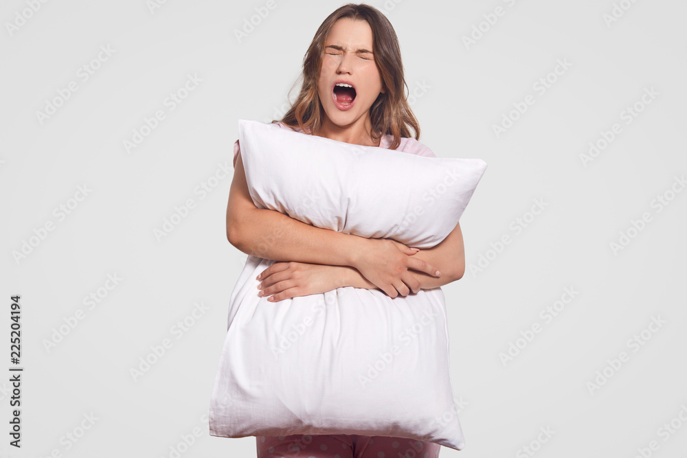 Annoyed desperate young woman yells loudly, keeps mouth widely opened, holds white pillow, poses against studio background, dressed in pyjamas. People, negative emotions and feelings concept Annoyed desperate young woman yells loudly, keeps mouth widely opened, holds white pillow, poses against studio background, dressed in pyjamas. People, negative emotions and feelings concept