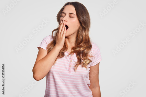 Indoor shot of pretty woman yawns, holds hand on mouth, dressed in casual clothes, keeps eyes shut, wants to have rest and sleep, isolated over white background. People and bed time concept.