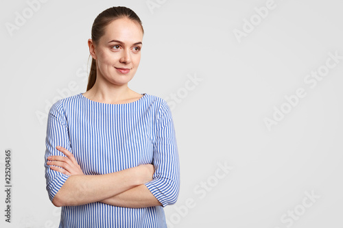 Horizontal shot of thoughtful European female with pony tail, keeps hands crossed, dressed in striped blouse, poses against white background with copy space aside for your promotion or advertisement