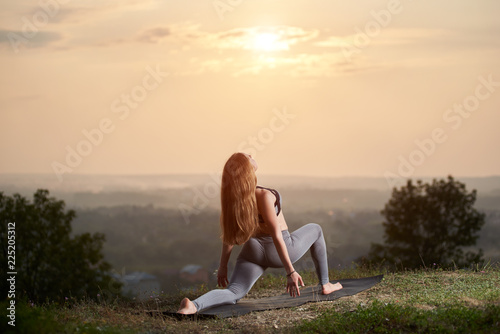 Young attractive long-haired athletic woman doing stretching yoga exercises on green hill on bright pink sky at sunset and foggy summer landscape background. Fitness and healthy lifestyle concept.