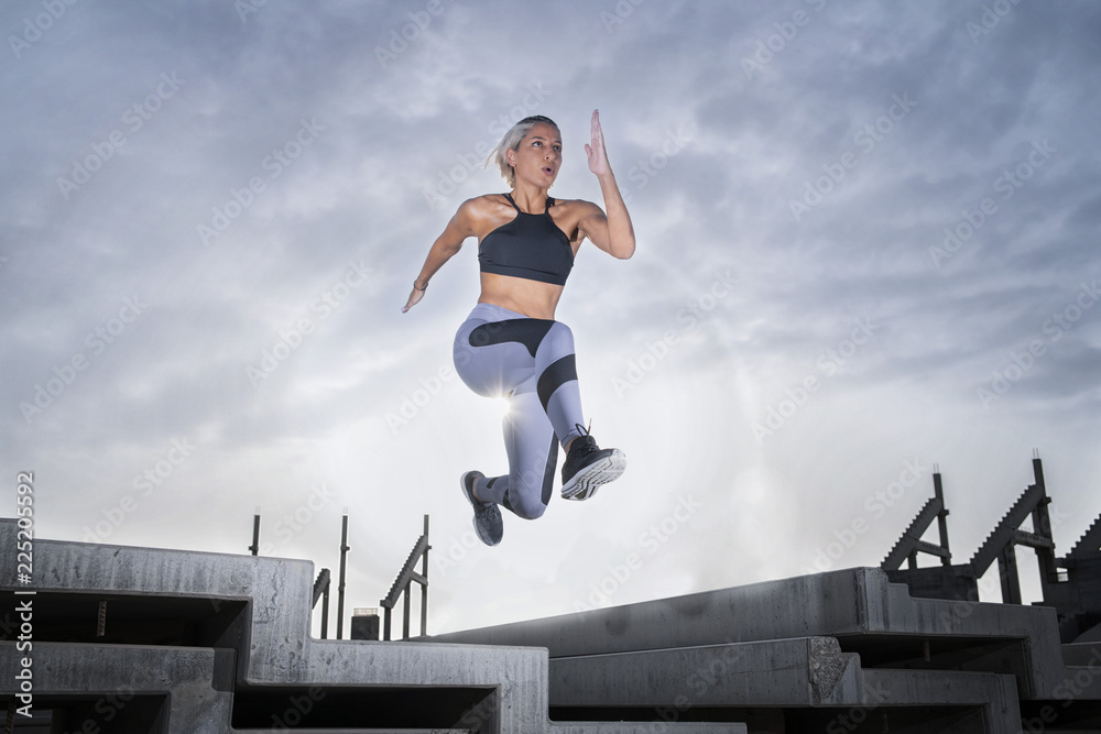 Middle Eastern Girl with short braided hair jumping of a stack of blocks on a construction site wearing gray and black fitness outfit on a hot bright sunny day. Middle Eastern Girl with short braided hair jumping of a stack of blocks on a construction site wearing gray and black fitness outfit on a hot bright sunny day.