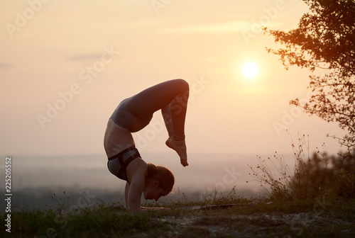 Silhouette of flexible woman in training outfit doing difficult complicated yoga exercises outdoors on background of clear light pink summer sky at sunset. Fitness, training and beauty concept.
