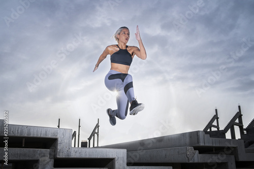 Middle Eastern Girl with short braided hair jumping of a stack of blocks on a construction site wearing gray and black fitness outfit on a hot bright sunny day. 