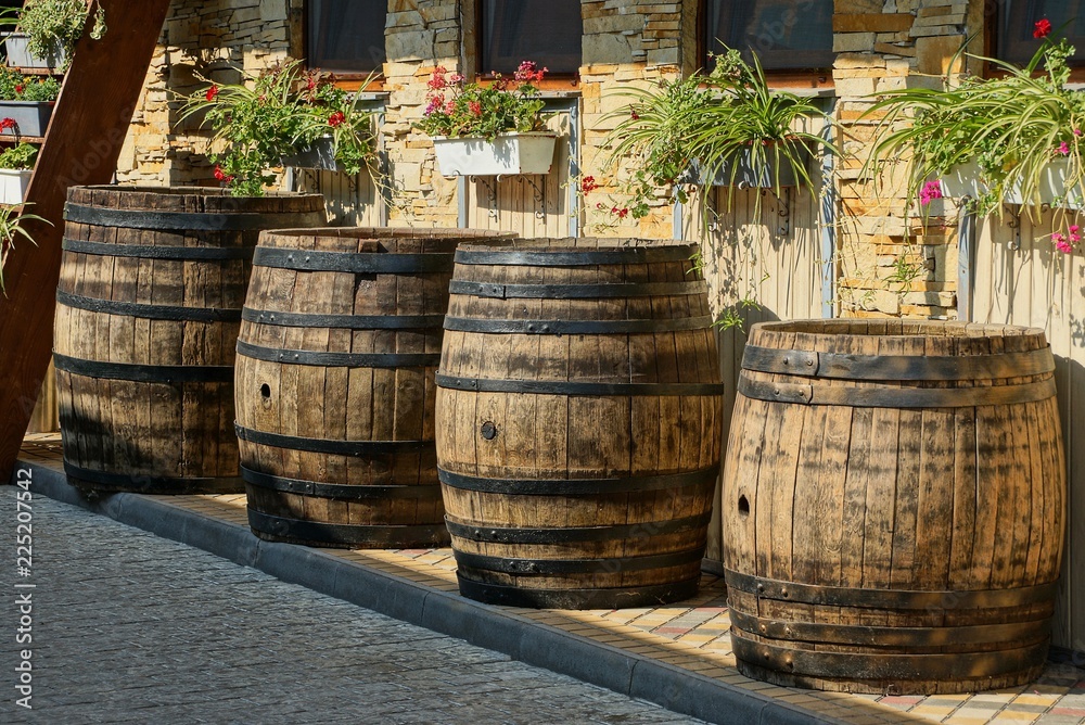 a row of brown wooden barrels stand on the sidewalk near the wall with flowerpots and windows a row of brown wooden barrels stand on the sidewalk near the wall with flowerpots and windows
