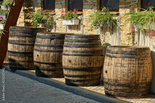 a row of brown wooden barrels stand on the sidewalk near the wall with flowerpots and windows © butus