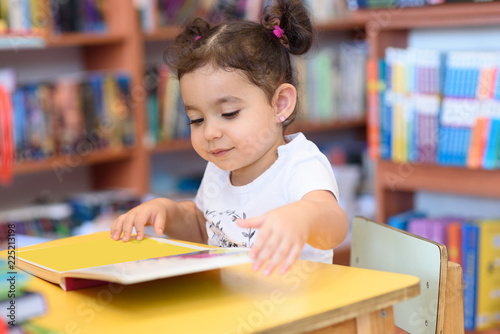 Little Girl Indoors In Front Of Books. Cute Young Toddler Sitting On A Chair Near Table and Reading Book. Child reads in a bookstore, surrounded by colorful books. Library, Shop, Shelving In Home.