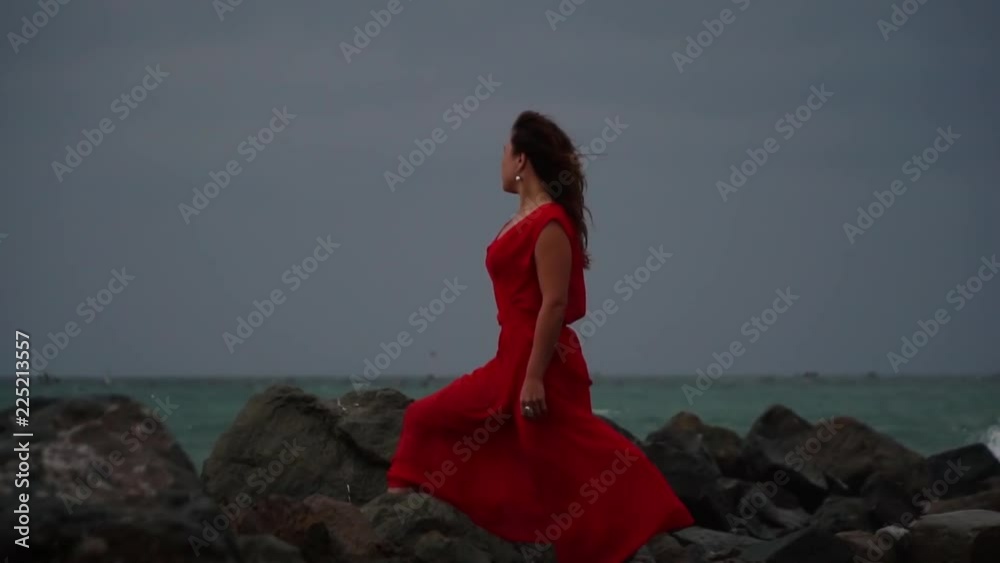 Girl in red dress stands in the raging sea on the rocks, Young woman near the sea during a storm. Girl in red dress stands in the raging sea on the rocks, Young woman near the sea during a storm.