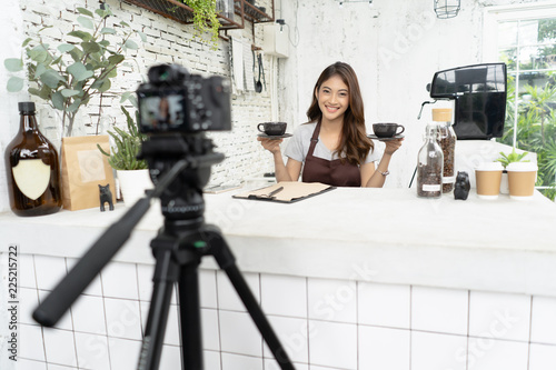 Attractive young Asian beautiful caucasian barista in apron smiling with recording video camera filming at  coffee shop counter. Startup Small Business Owner Concept. Making vlog concept. photo