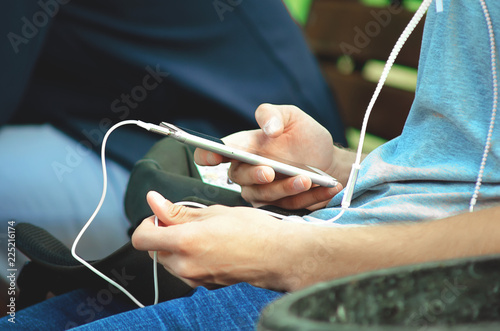 A young man sits on the street and uses a smartphone with headphones. Hands close-up.