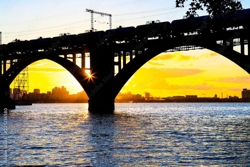 Naklejka premium Silhouette of a beautiful arched railway bridge and wagons on the Dnieper river at sunset. Dnipropetrovsk (Dnepr, Dnipro, Dnepropetrovsk) Ukraine.