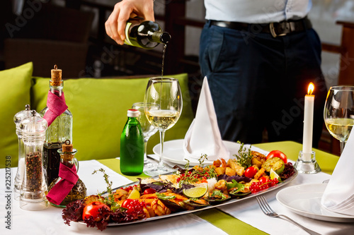 Elegant waiter is pouring wine to a glass at decorated with candle table with meat set dish.